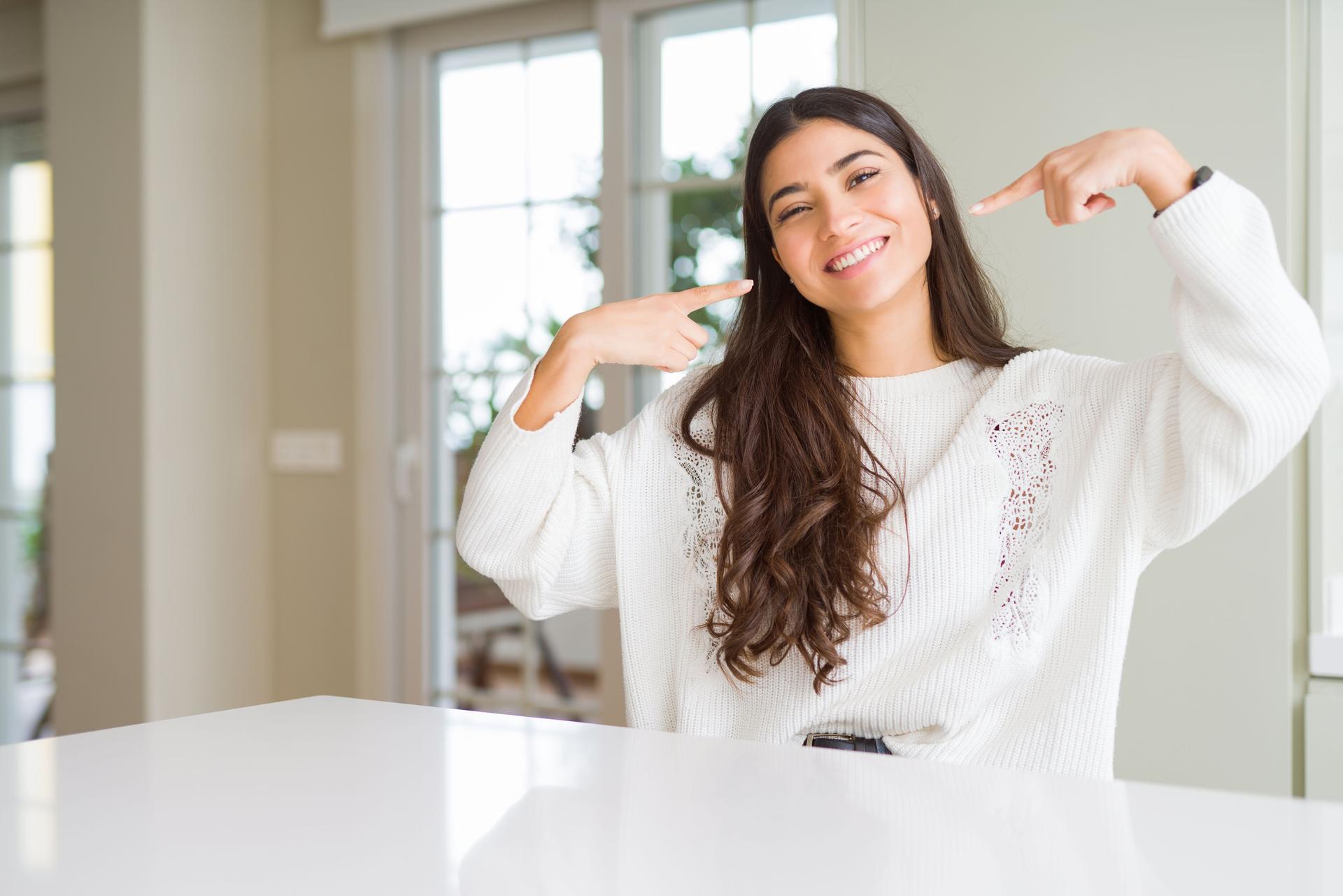 Young beautiful woman at home on white table smiling confident showing and pointing with fingers teeth and mouth. Health concept.